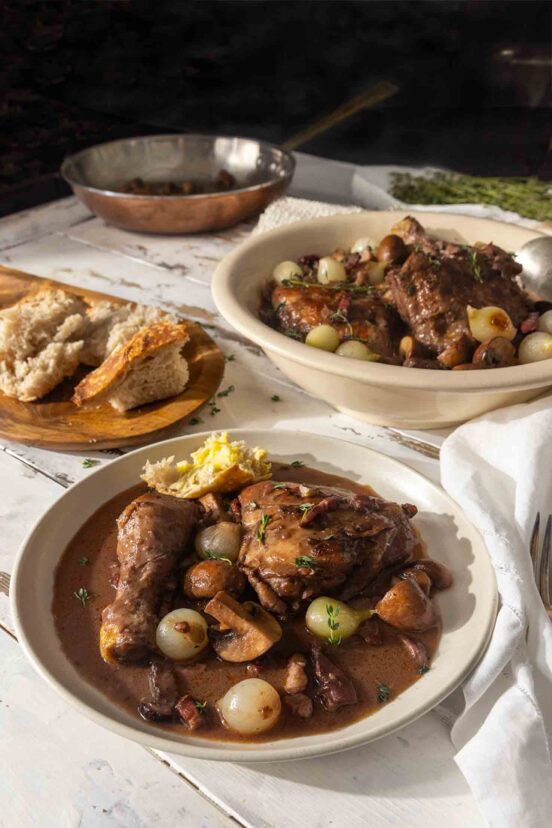 Two bowls of coq au vin on a table with sliced bread and fresh herbs.