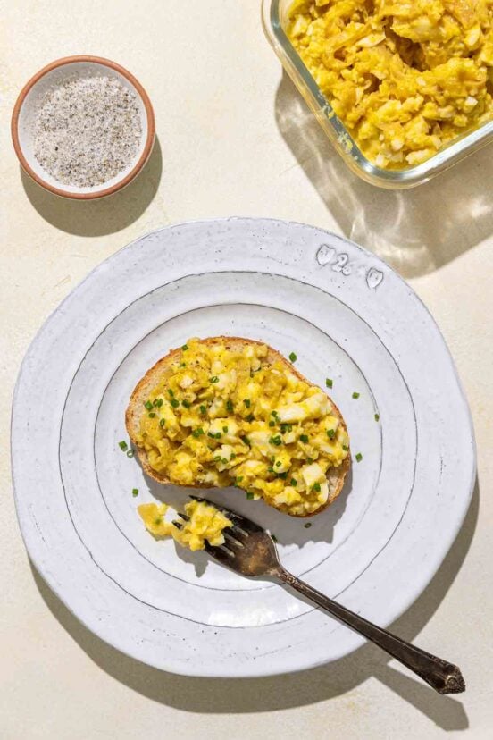 An open-faced egg salad sandwich topped with minced chives on a white plate with a bowl of salad and a dish of salt and pepper nearby.