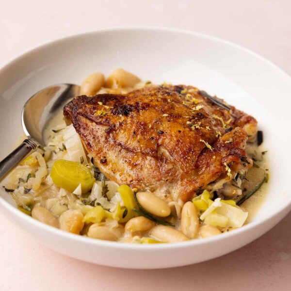 A bowl with one crispy chicken thigh on top of a mixture of leeks and cannellini beans.