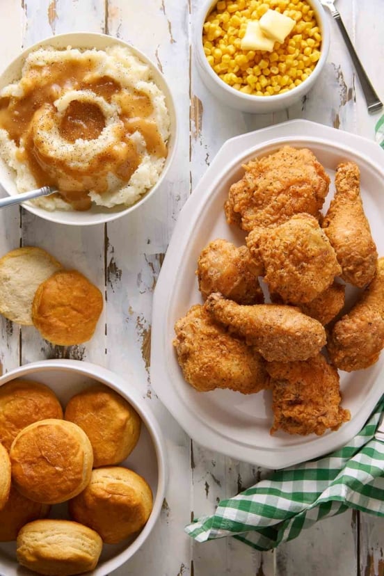 Pieces of batter-fried chicken piled on a white platter, along with biscuits, mashed potatoes, and buttered corn.