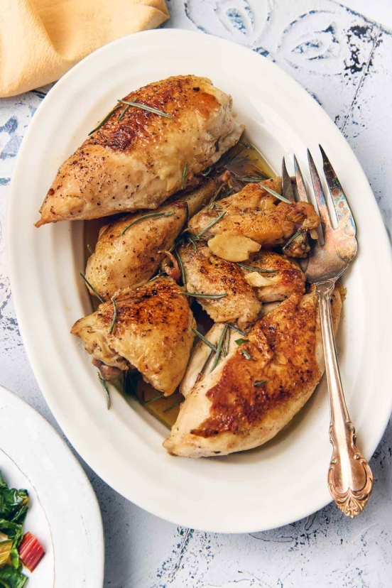 Pieces of browned chicken topped with garlic and rosemary leaves on a white oval platter.