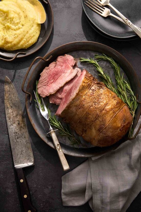 A partially sliced boneless leg of lamb on a platter with rosemary sprigs around it.