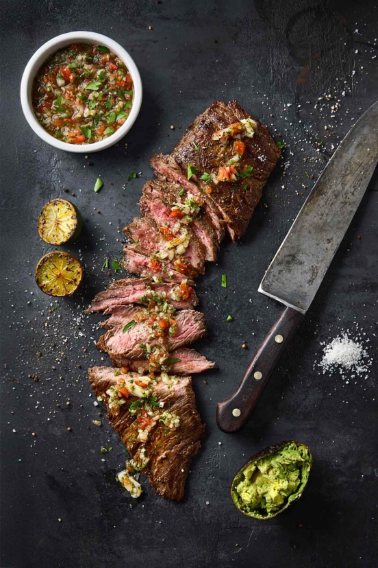 Sliced skirt steak topped with salsa with a bowl of salsa and charred lime halves on the side.