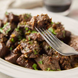 A fork holding a single garlic butter steak bite with a platter of steak bites nearby.