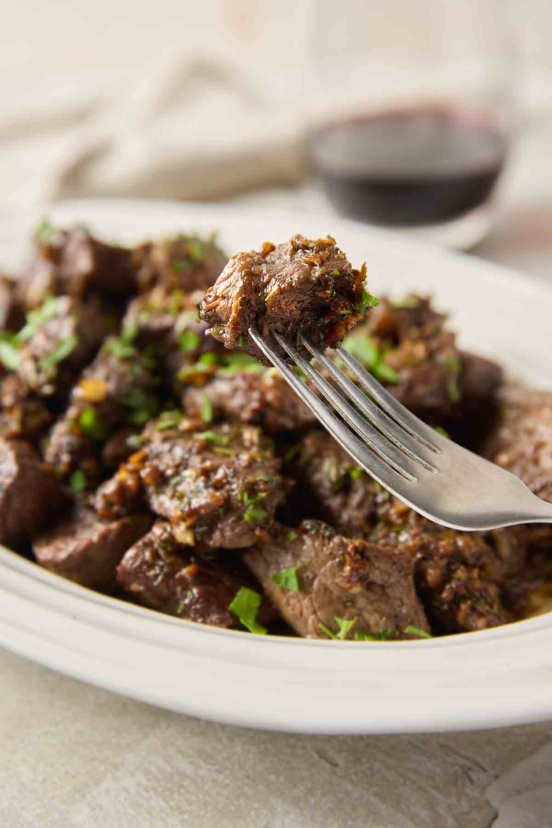 A fork holding a single garlic butter steak bite with a platter of steak bites nearby.