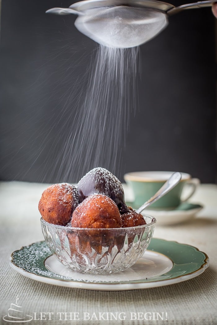 Donuts topped with chocolate ganache and powdered sugar in a glass bowl on a green and white decorative plate.