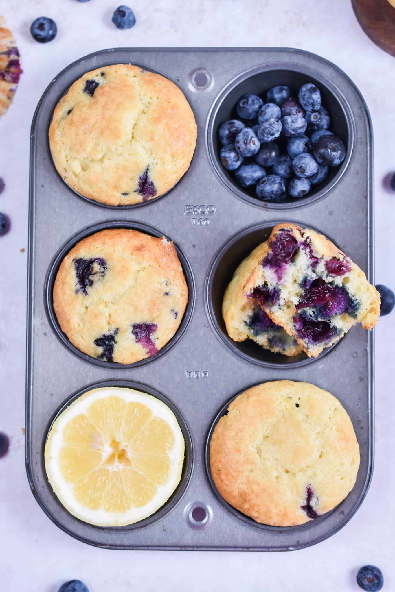 An overhead shot of the blueberry muffins with sour cream in a muffin tin with one muffin cup holding blueberries and one holding a half of a lemon.