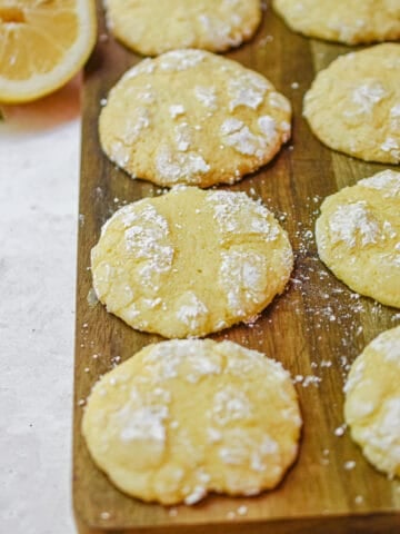 The 3 ingredient lemon cookies are laid out on a cutting board with a cut lemon in the background.