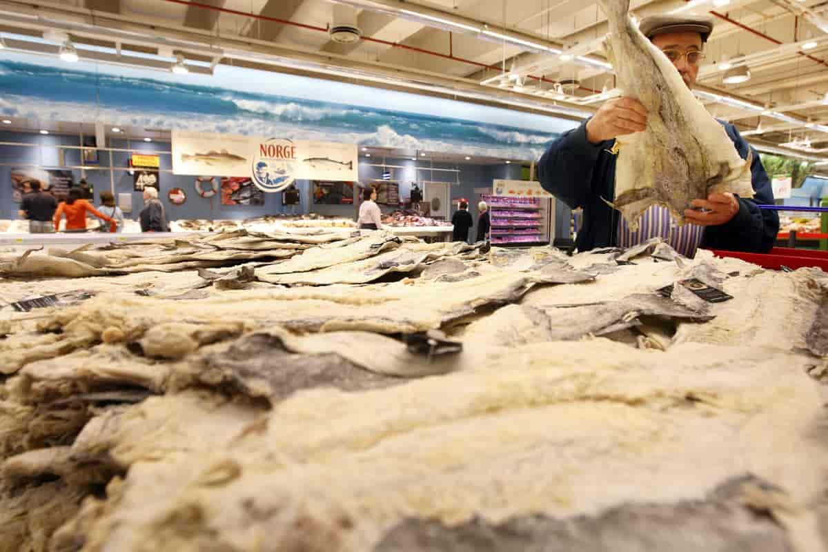 Inne i et supermarked. En mann med sixpence og briller holder opp en stor tørket fisk. Fisken skjuler nesten hele mannens overkropp og hode. I forgrunnen ligger mange flere tørka fisk stablet. Foto