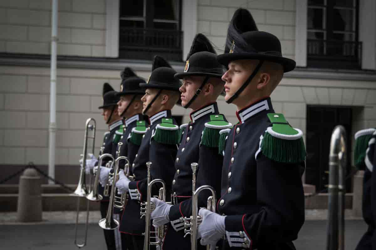 Foto av fem gardister på rekke. Alle har svart uniform og svart hatt med svart dusk. Fire av dem holder hver sin trompet, den femte holder en trombone. 