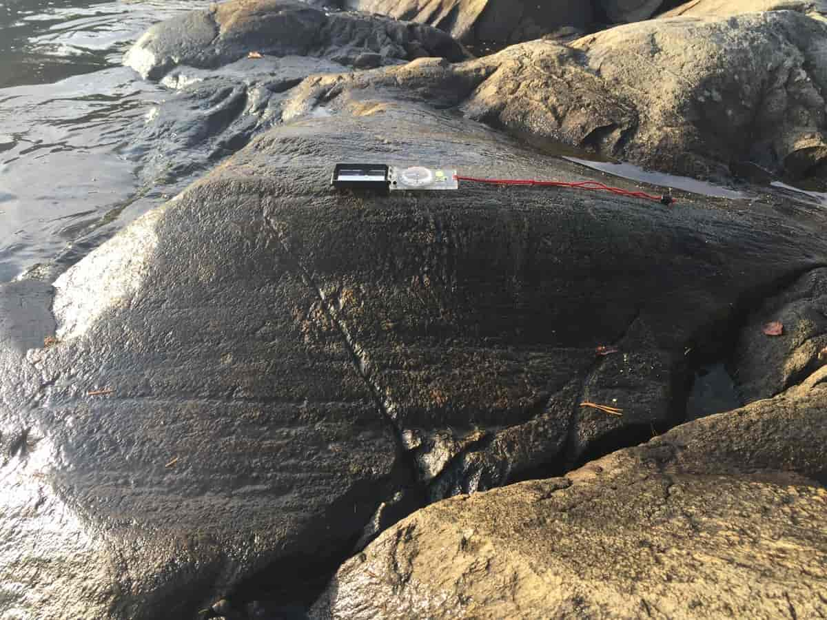 Foto av svaberg med noen horisontale fordypninger i striper. Et kompass med rød snor ligger på en av steinene for å vise størrelsen og retningen på stripene. De er på linje ut mot vannet.