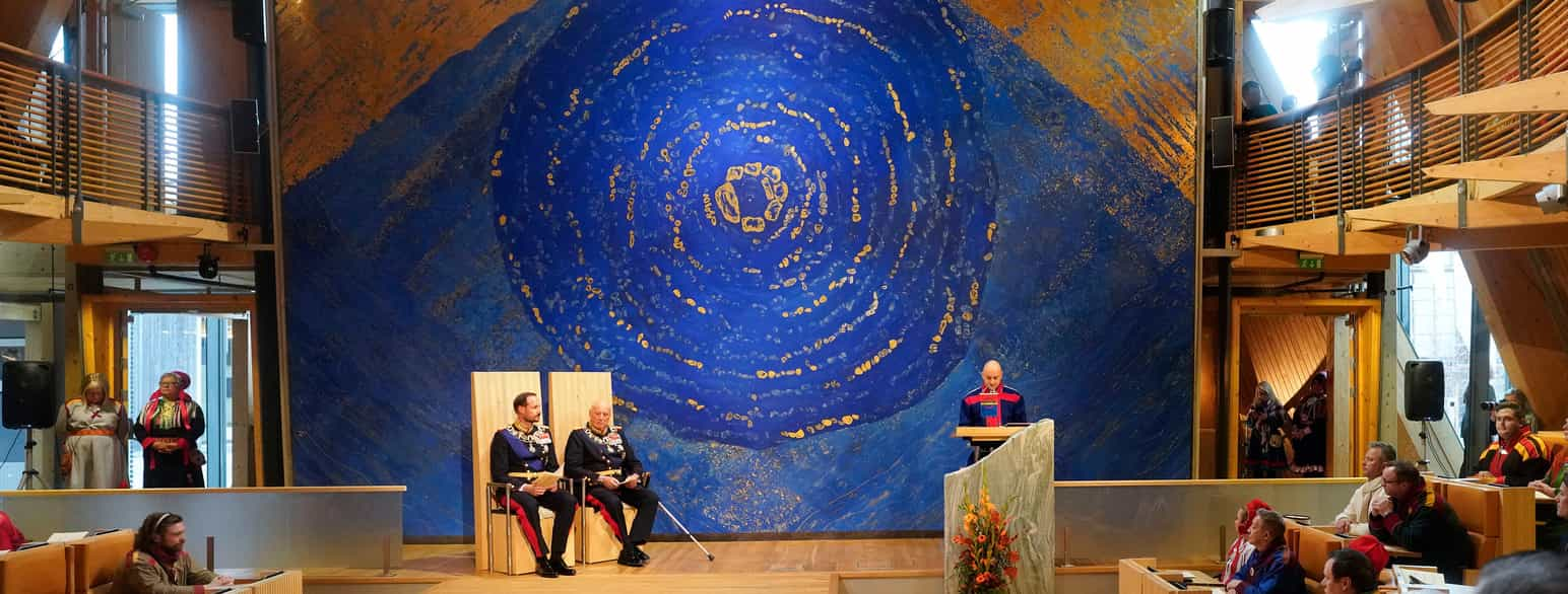 En stor sal med et podium. På venstre side av podiumet sitter to menn i paradeuniform på hver sin stol med høy rygg. På høyre side står en mann bak en talerstol med et lite samisk flagg på. Bak podiet er et stort kunstverk: en blå sirkel med uregelmessige striper i gull dekker nesten hele veggen. Foto