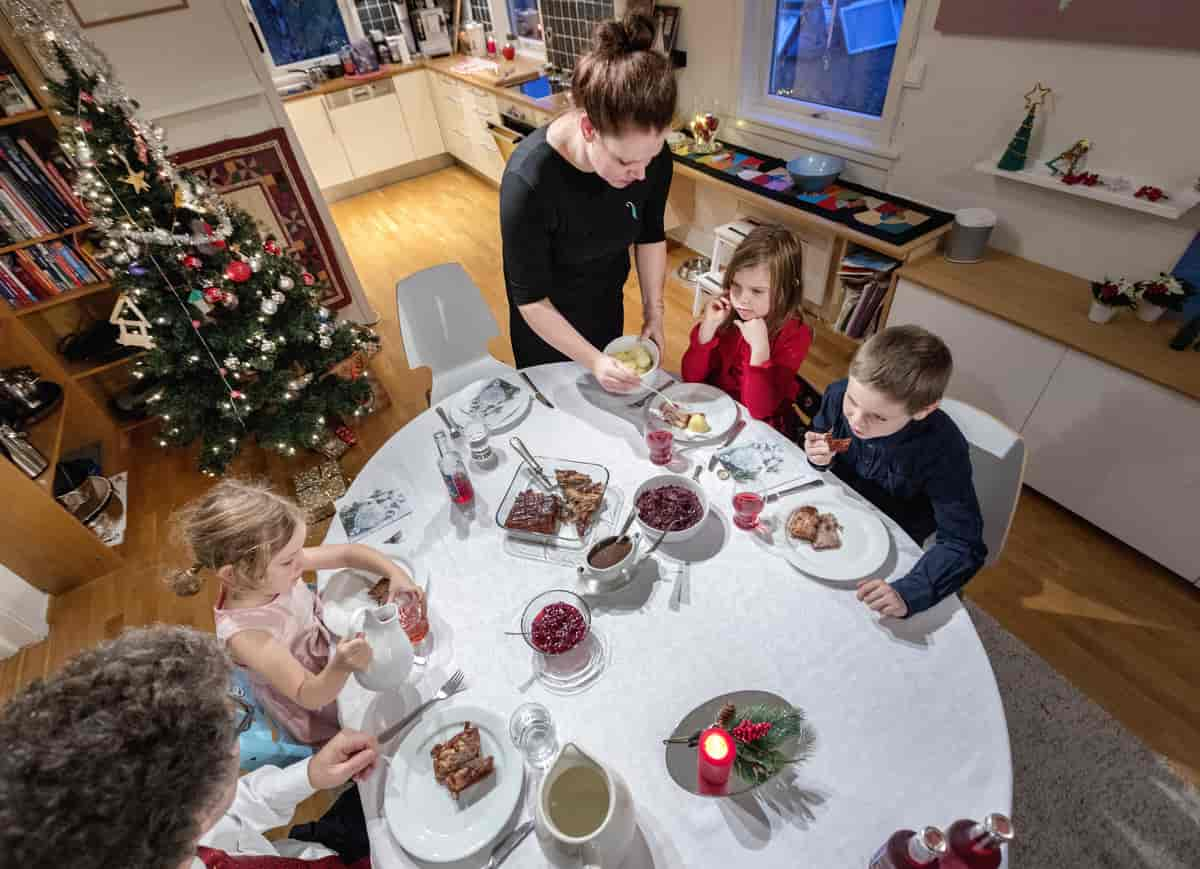 En familie som sitter rundt et bord. Alle er pyntet. Det er dekket med hvit duk og på bordet er det ribbe, poteter, rødkål. I bakgrunnen står det et juletre.