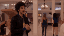 a man in a black leather jacket stands in a room with a chandelier