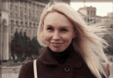 a woman with blonde hair is smiling and looking at the camera while her hair blows in the wind .