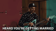 a man playing a guitar and singing into a microphone with the words heard you 're getting married behind him
