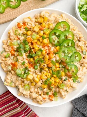 corn dip with cream cheese topped with corn, green onions, and jalapenos, and hot sauce in a white bowl