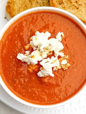 red pepper and gouda soup topped with popcorn in a white bowl with toasted bread on the side.