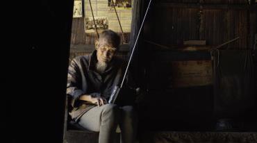 Elderly person holding a radio, seated inside a dim wooden cabin.