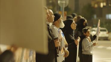 A diverse group of people standing in line, some wearing hijabs, holding posters. 