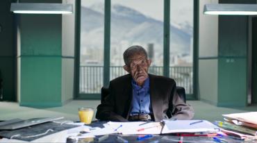 Exhausted elderly man sits at a cluttered, high rise office desk.