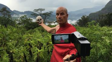 Man holding a skull in a forest; camera in foreground capturing him in a robe garb. 