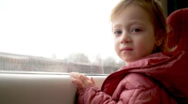 Young child in a pink jacket in car turning from window at camera.