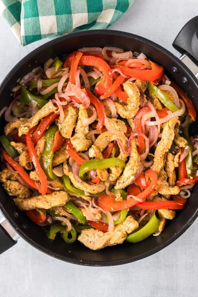 A skillet containing cooked strips of chicken, red and green bell peppers, and onions for chicken fajita wraps.