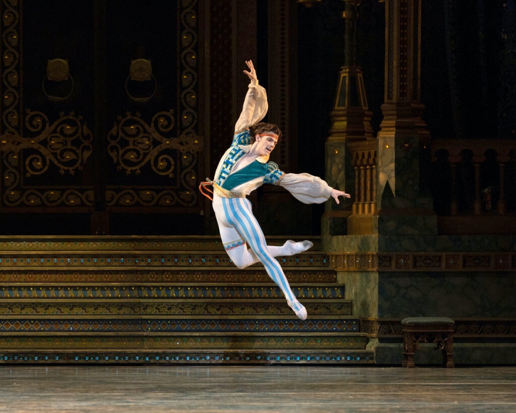 Jake Roxander, wearing white and blue striped tights, red headband, and a white and blue tunic with billowing sleeves, jumps up into the air and pikes his body to his left with his right leg tucked slightly behind him. He reaches left with his upper body and looking towards his right foot. He smiles energetically. He performs onstage in front of an elaborate ballroom set.