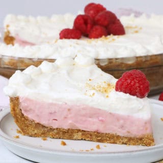 A slice of raspberry cream cheese pie with graham cracker crust topped with homemade whipped cream and a fresh raspberry on a white plate.