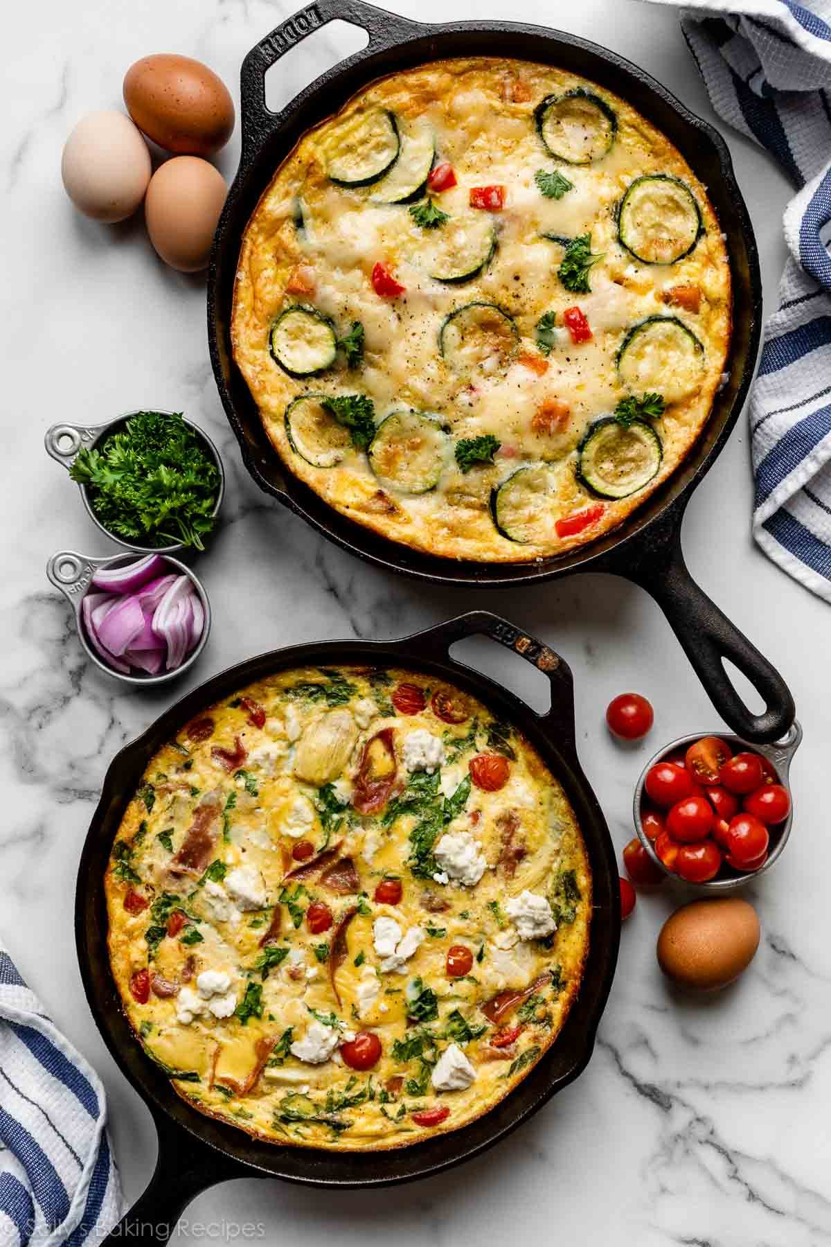 two frittatas in cast iron skillets on marble counter with tomatoes, red onions, chopped parsley, and eggs next to them.