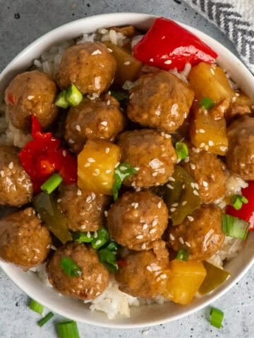 Slow cooker teriyaki meatballs over a bowl of rice on the table with a white and black tea towel to the side.