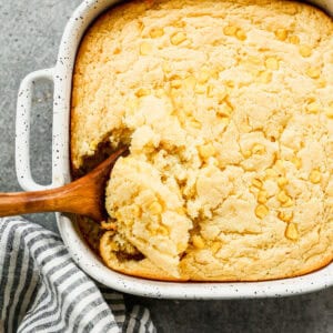 A pan of the best corn casserole baked in a 9x9 square pan and with a wooden spoon scooping some up to serve.