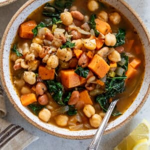 a bowl of soup filled with vegetables, potatoes and beans beside a cut lemon wedge