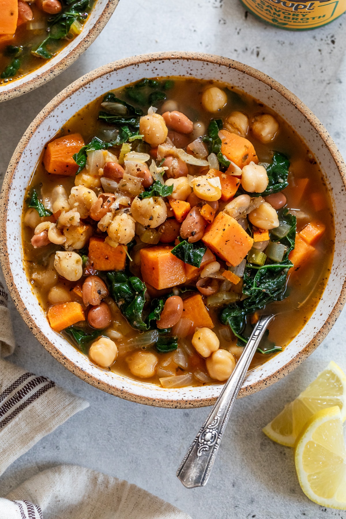 a bowl of soup filled with vegetables, potatoes and beans beside a cut lemon wedge