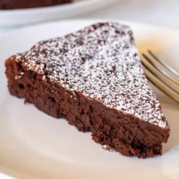 Piece of bittersweet chocolate cake with powdered sugar on white plate with fork.