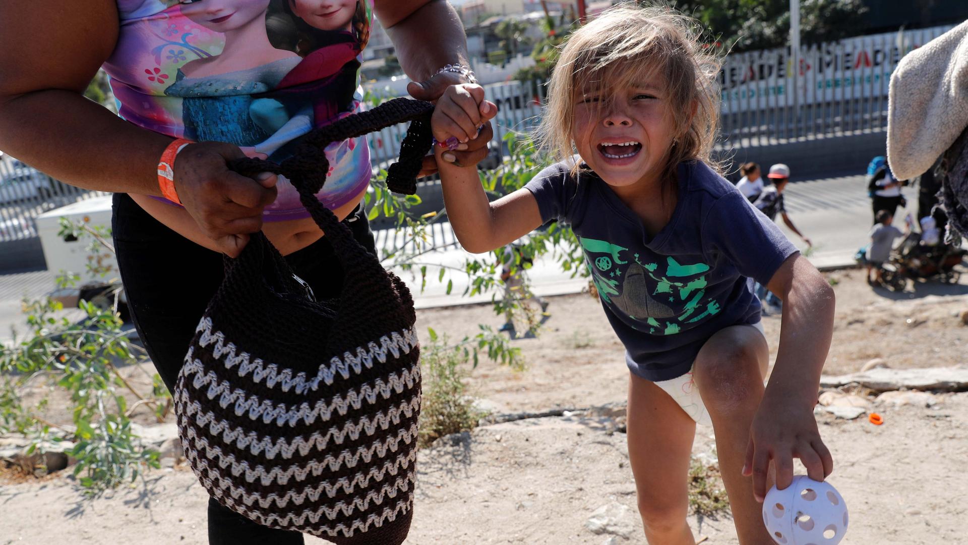 A small girl in a t-shirt with bare legs and feet cries she and her mother run up a dirt hill.