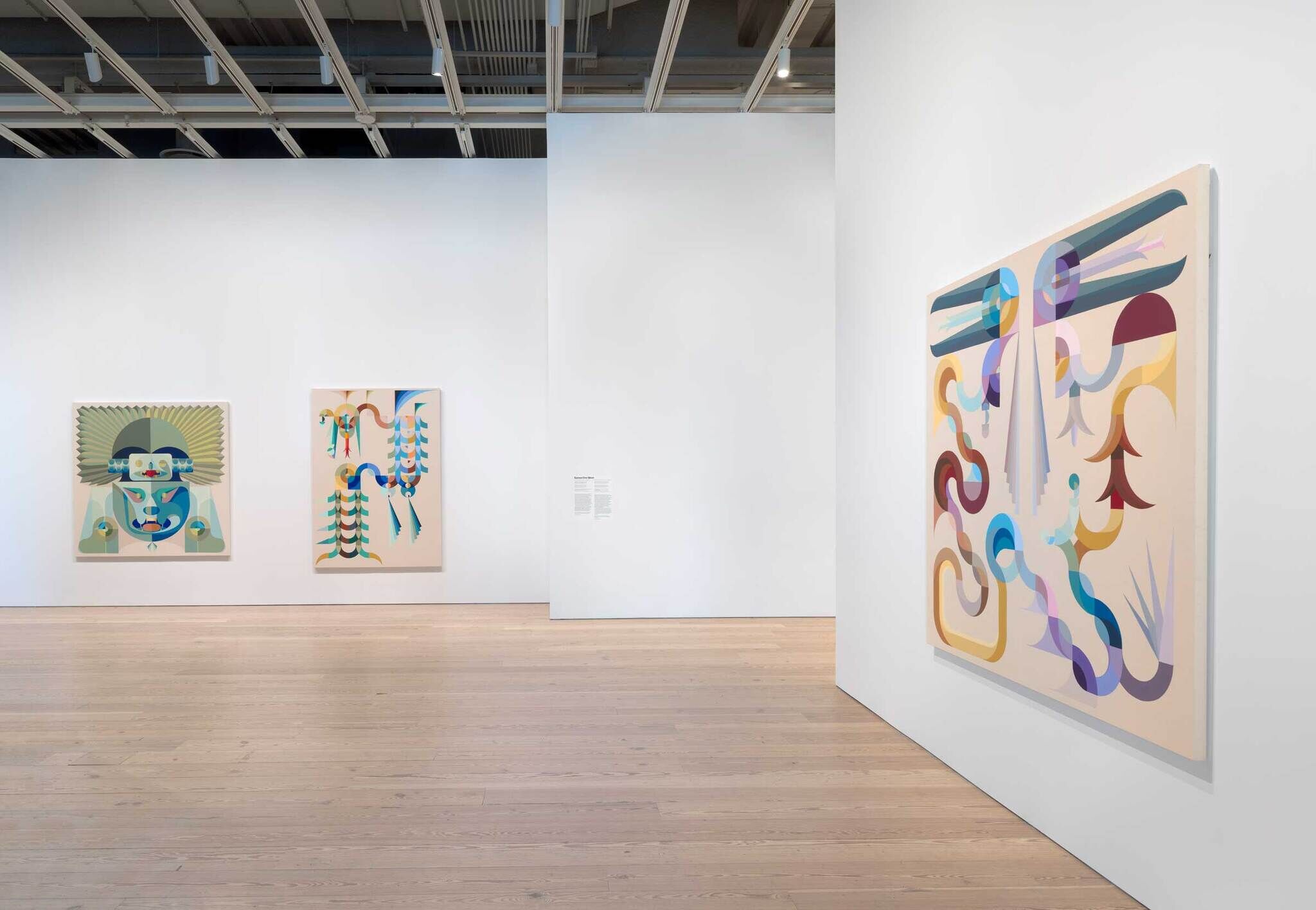 A gallery at the Whitney Museum featuring Eamon Ore-Giron's paintings. The space has light wooden floors and white walls, with three large paintings on display. The painting closest to the viewer on the right wall showcases abstract, flowing shapes in pastel colors, including purple, blue, and yellow. In the background, two other paintings are visible: one featuring a detailed face with geometric patterns and the other with vibrant, abstract forms similar to the painting on the right. The gallery has a clean, modern aesthetic with an open layout and high ceilings.