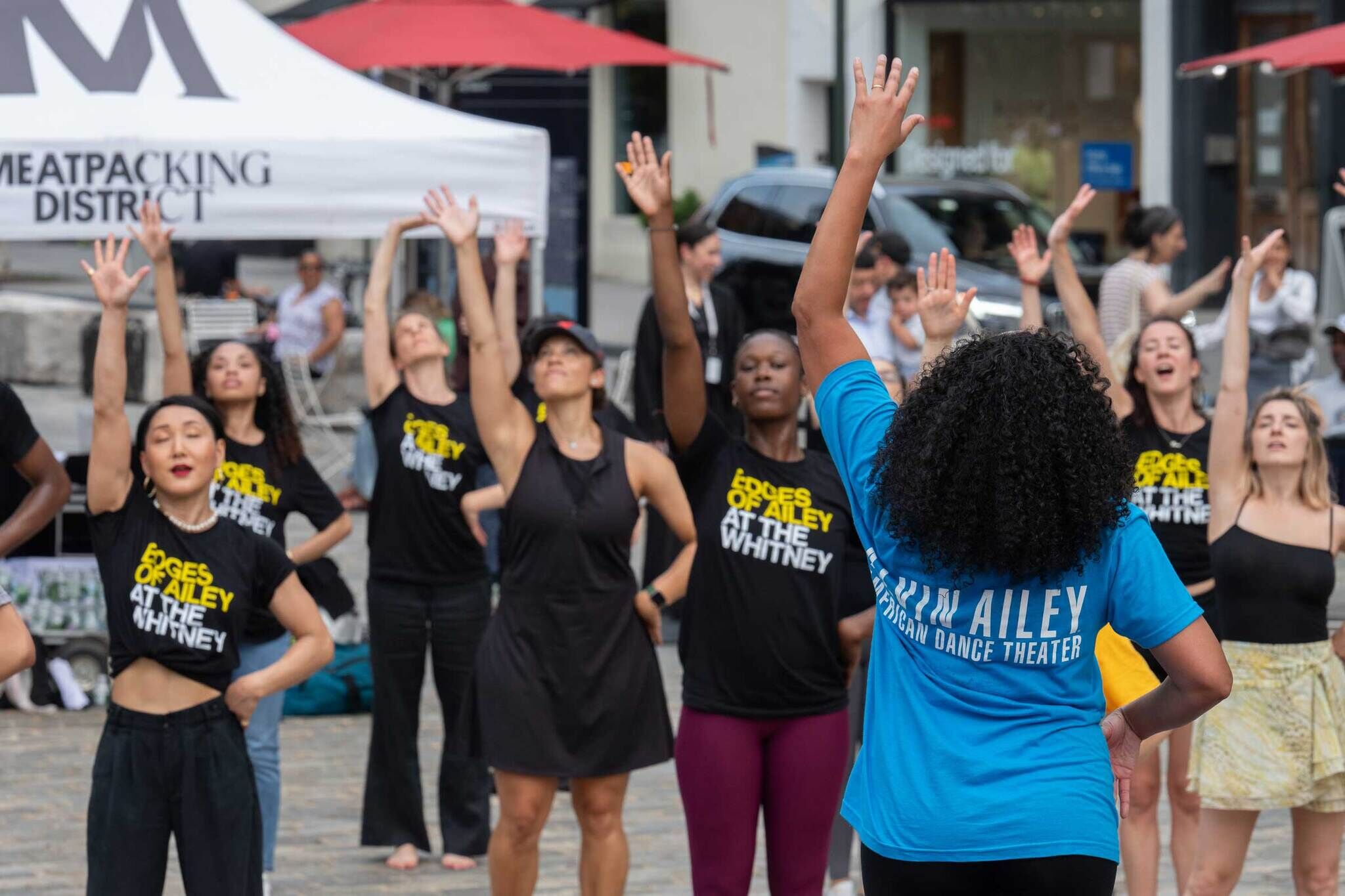 A group of people in a public space participate in a dance class, led by an instructor in a blue shirt. They raise their arms in unison.