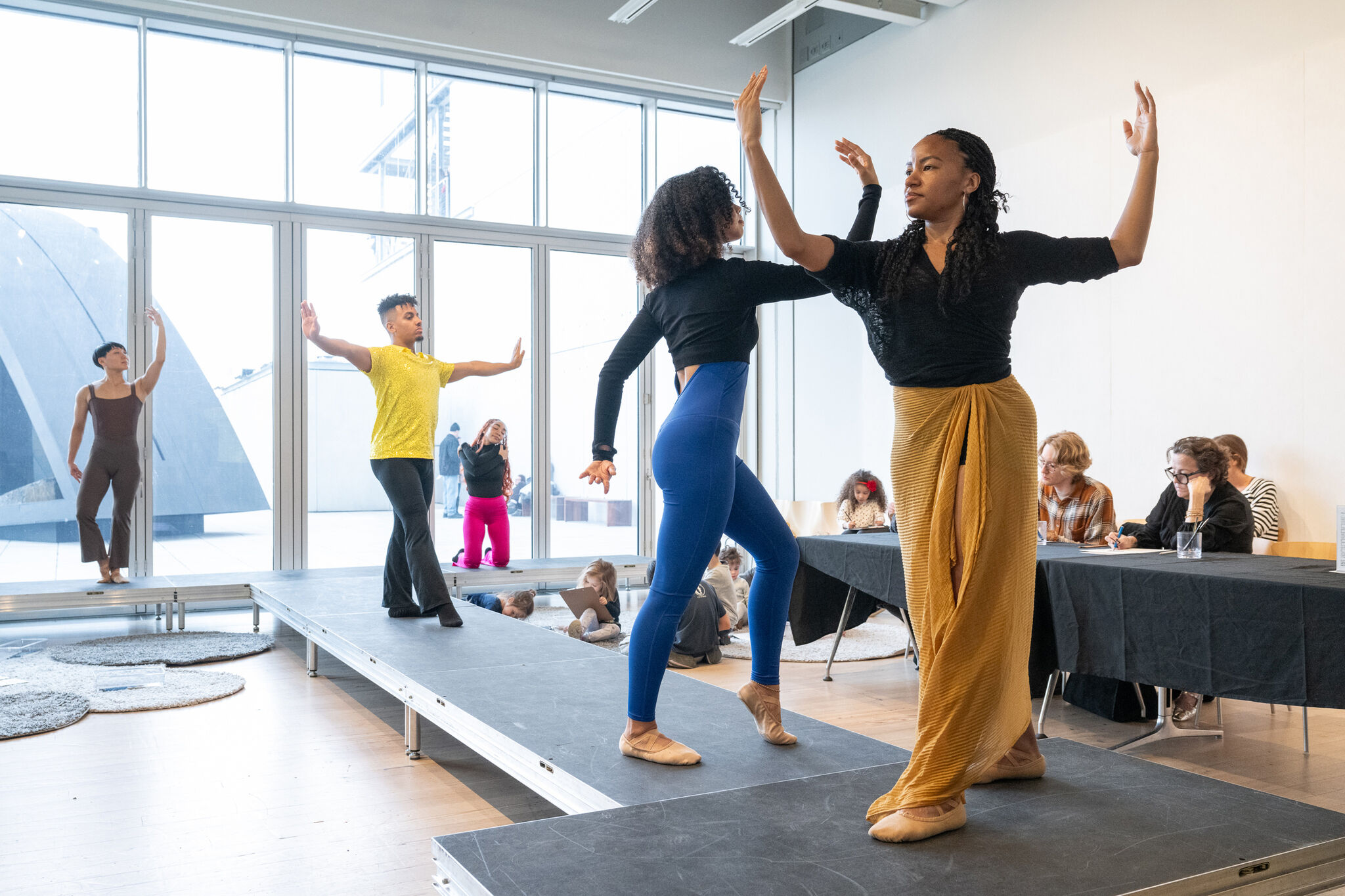 A figure drawing session at the Whitney Museum during a Free Second Sunday event. Dancers pose gracefully on an elevated stage, holding expressive and dynamic stances for attendees to sketch. Participants of all ages, seated at tables and on the floor, are immersed in capturing the dancers' forms. The space is bright and open, with large windows offering natural light and a view of the museum’s outdoor surroundings, creating a vibrant and creative atmosphere.