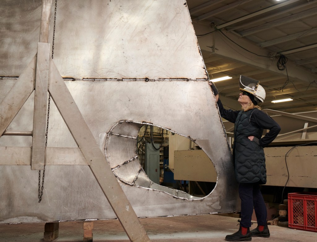 A white woman in a black padded jacket and dark pants stands with hands on hips in front of a large, metallic, geometric-shaped sculpture in an industrial workshop setting. The sculpture has rough surfaces and an open circular area. The environment includes visible machinery and the ceiling is supported by beams.