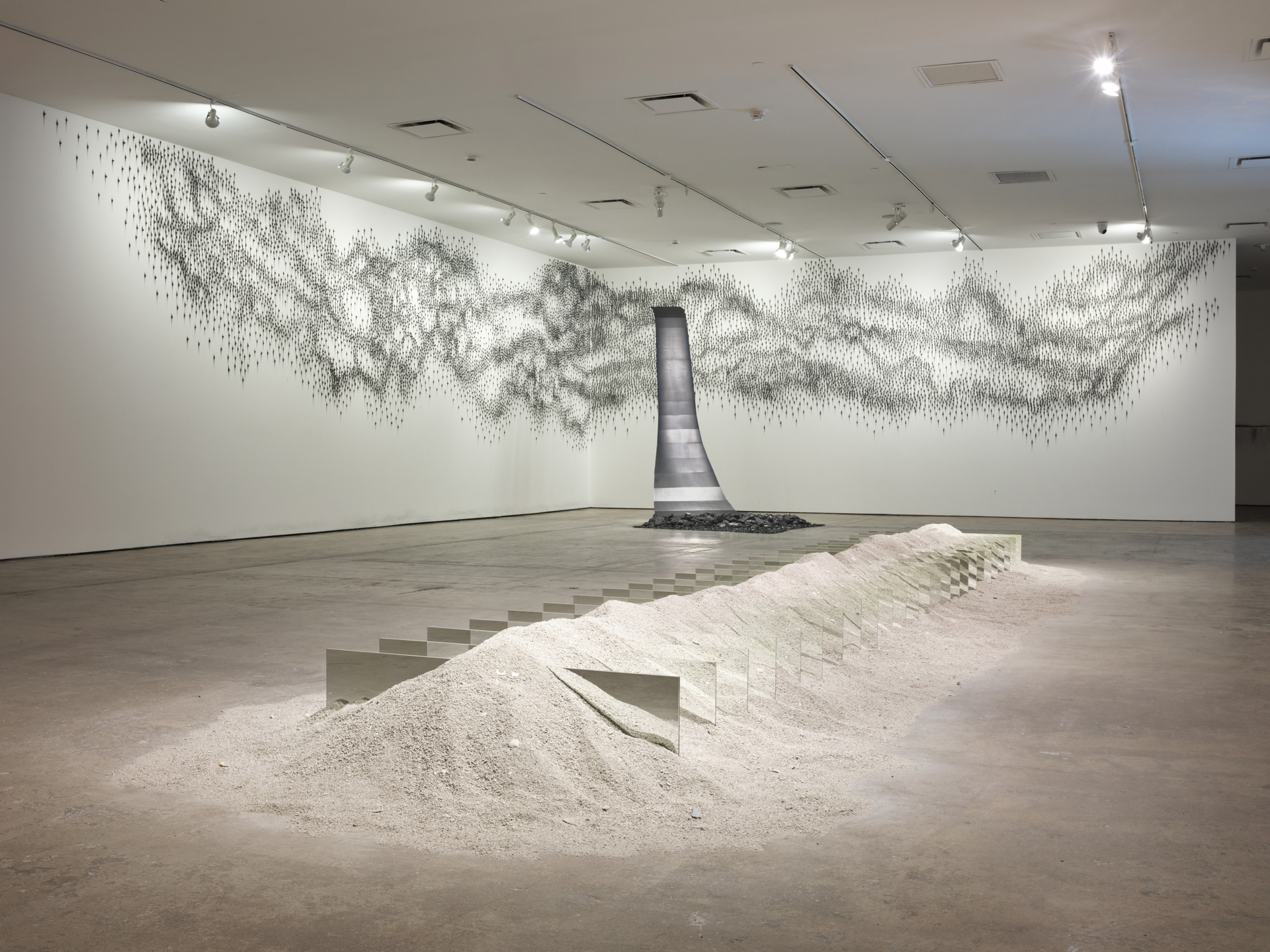 A cascading silver form in the corner of a gallery is shown amid grphite marks covering a wall. In the foreground, a linear lump of sand is punctuated with vertical rectangular mirrors.