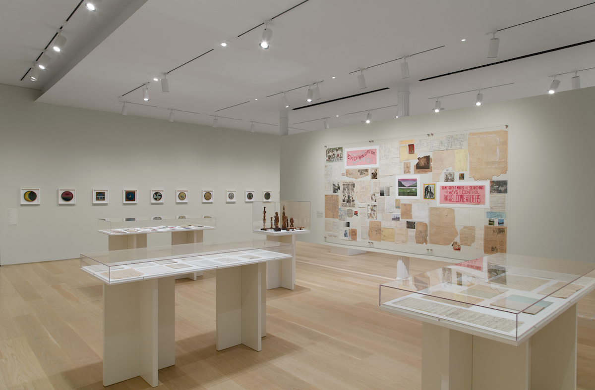 A gallery with vitrines in its center and drawings on its walls.