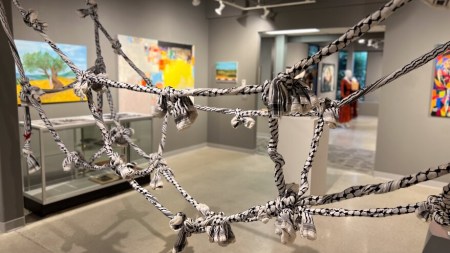 A net of knotted kaffiyahs is in the foreground of an image of a gray gallery. Colorful but out-of-focus paintings can be seen in the background.