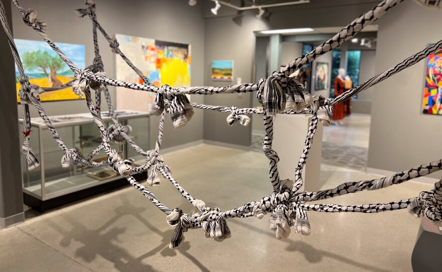 A net of knotted kaffiyahs is in the foreground of an image of a gray gallery. Colorful but out-of-focus paintings can be seen in the background.