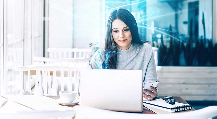 Frau sitzt in einem Café und arbeitet an ihrem Laptop, während sie ihr Smartphone in der Hand hält