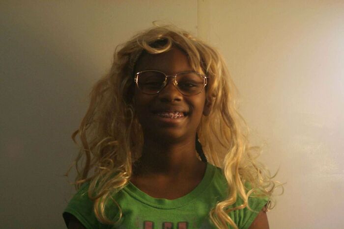 Child in glasses and braces, wearing a blonde wig, smiling joyfully in a casual setting, embodying blunder years.