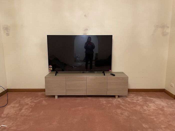 Modern TV on a wooden stand in a sparsely furnished room, reflecting the silhouette of a person taking a photo.