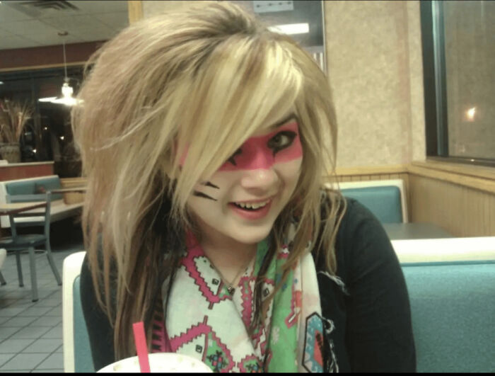 Young person with an emo hairstyle and colorful face paint, sitting in a diner, embodying blunder years fashion.