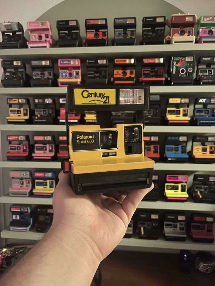 Hand holding a yellow Polaroid Spirit 600 camera, with a backdrop of colorful vintage camera collections.